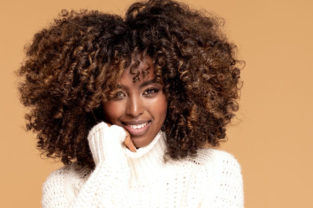 Happy beautiful African girl with afro hairstyle posing in cozy sweater on beige studio background.